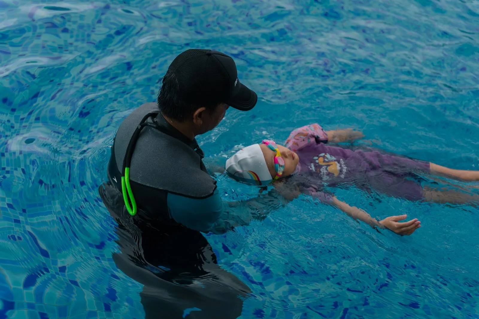 A coach in a black rash guard supporting a child floating on her back