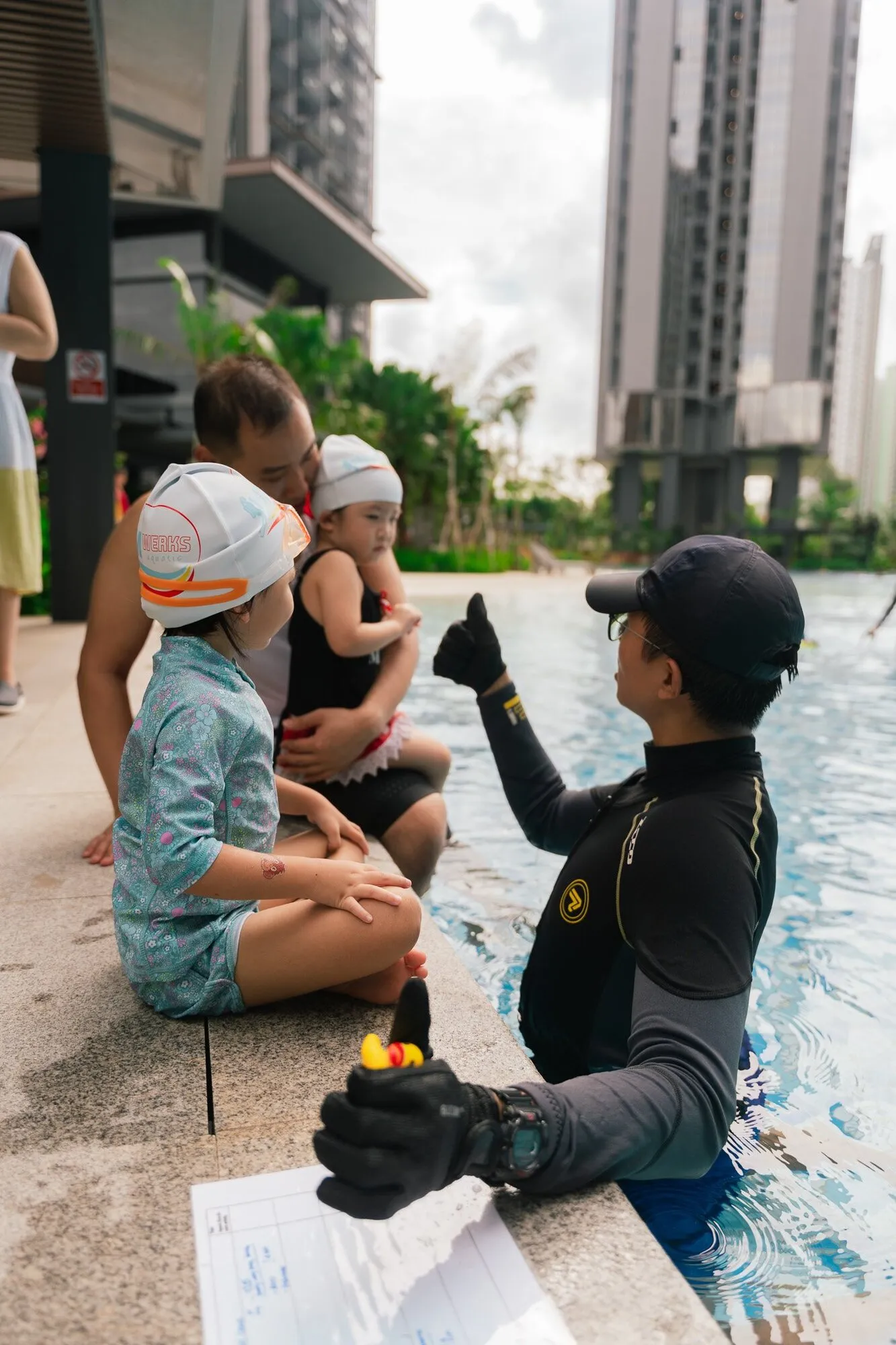 A trainee coach at the pool edge giving a thumbs-up to young swimmers in caps